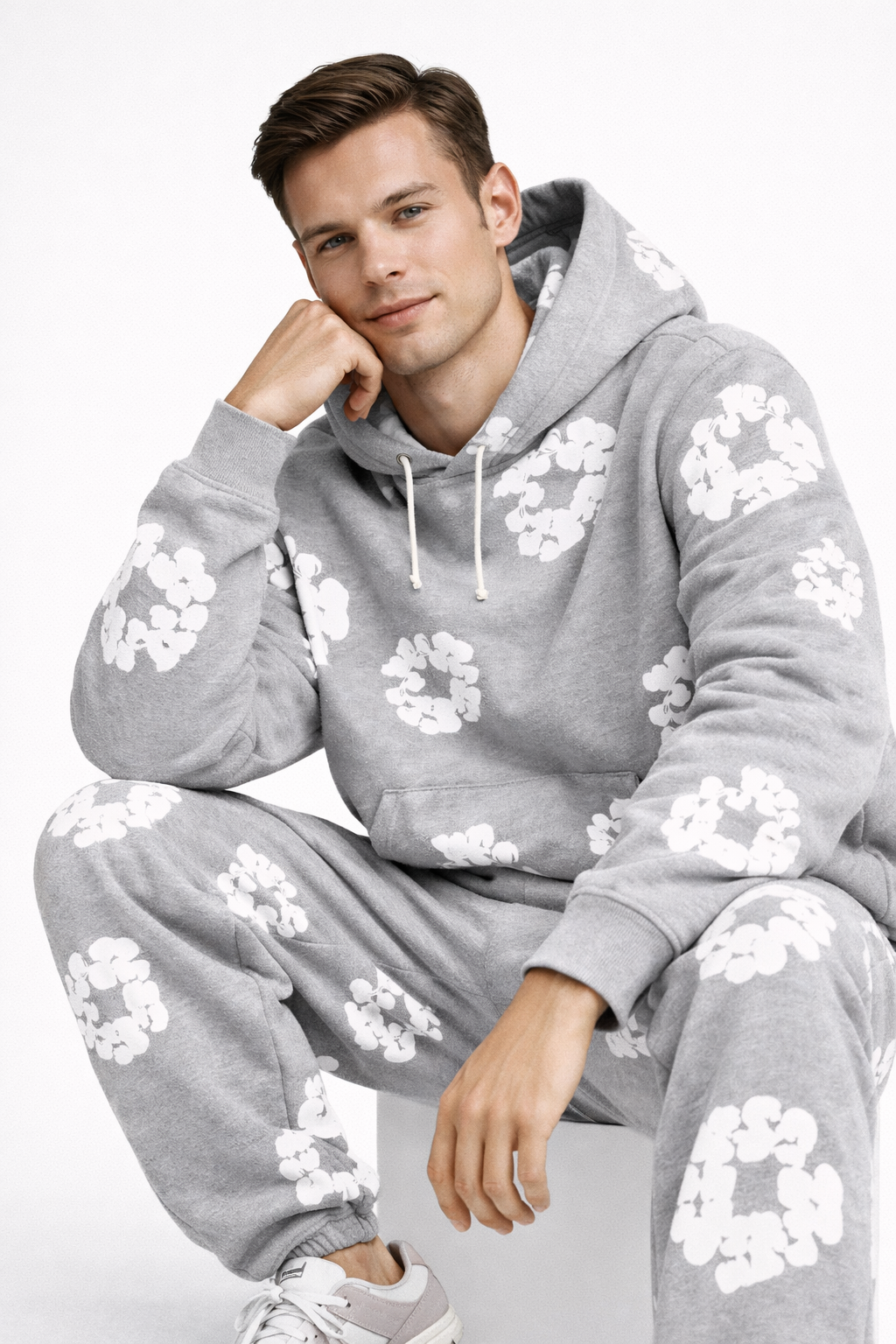 Ung mand iført grå hættetrøje og joggingbukser med hvide blomsterprint, sidder afslappet. Moderne streetwear, komfortabelt tøj, casual stil.