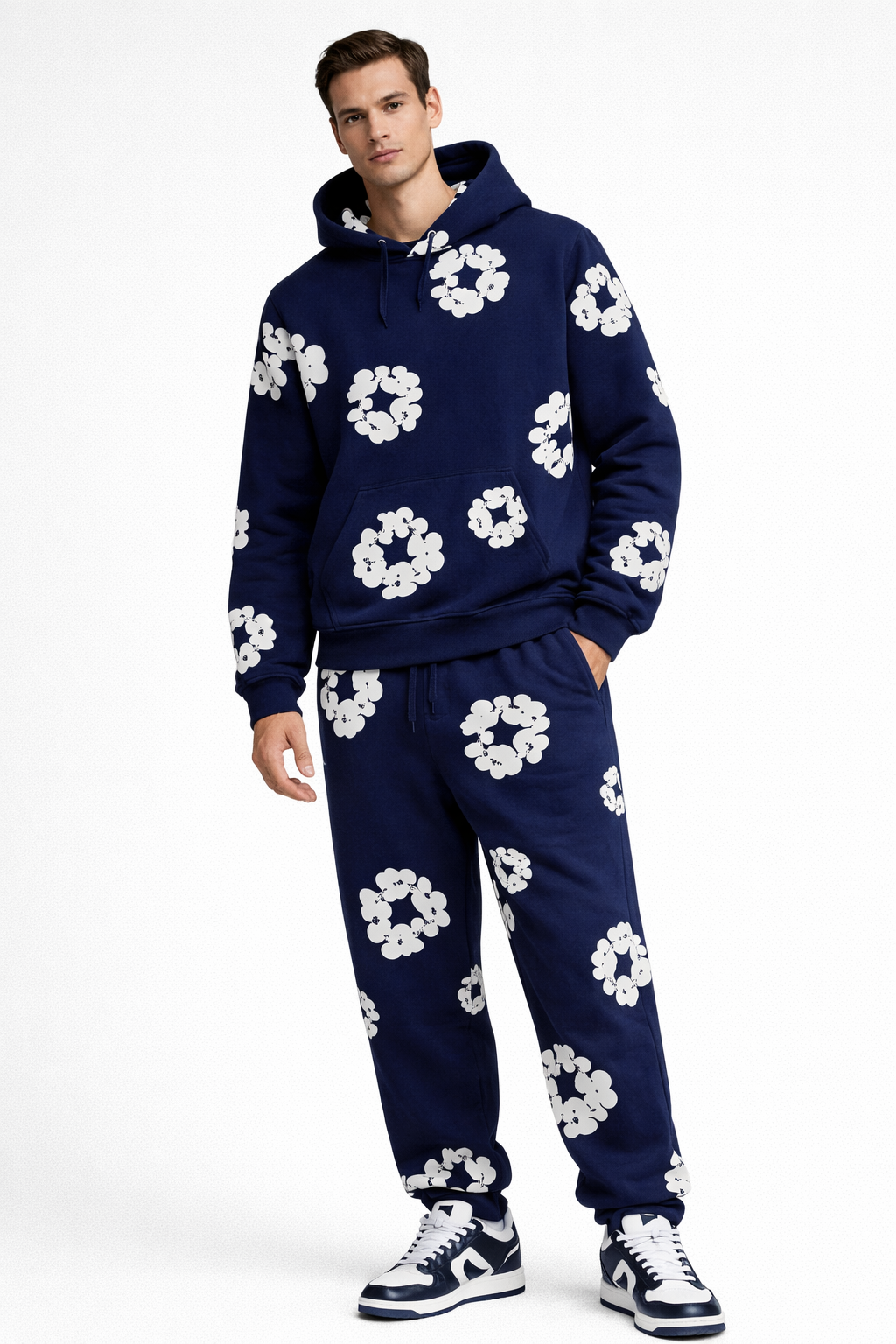 Mand iført mørkeblå hættetrøje og joggingbukser med hvide blomsterprint, afslappet stil, moderne streetwear, sneakers, hvid baggrund.
