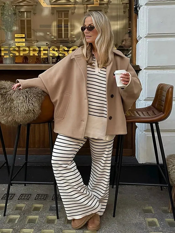 Kvinde i stribet tøj og beige frakke, holder kaffekop foran café. Mode, afslappet stil, solbriller, efterårstøj, byliv.