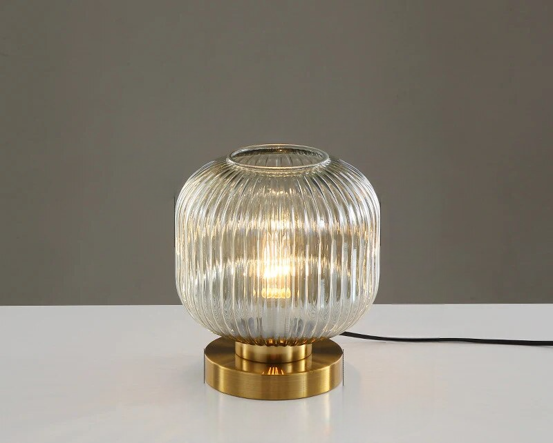 Glasbordlampe med rillet skærm og gylden base på hvidt bord. Moderne design, ideel til stueindretning og belysning.