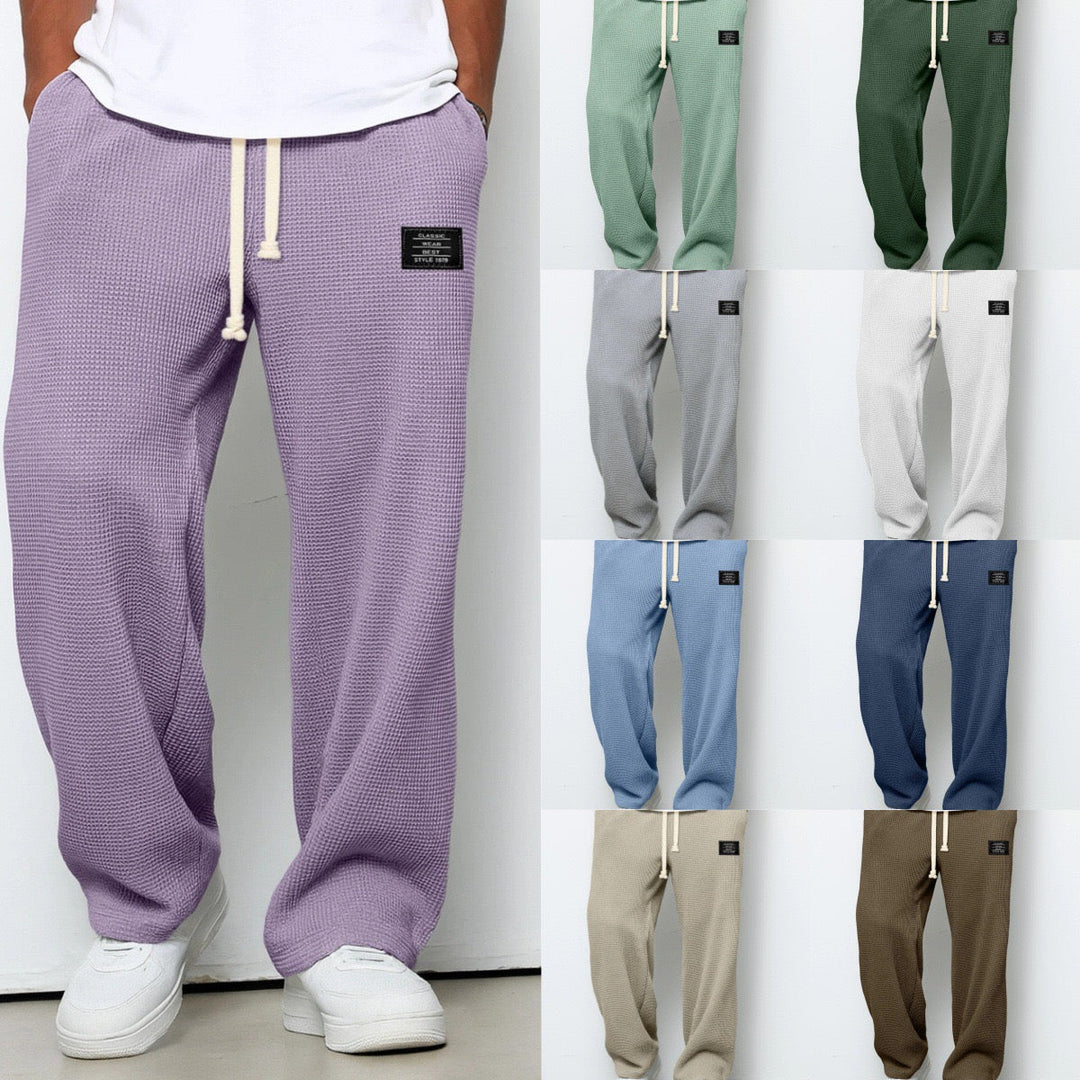 Mand iført lilla sweatpants med snor, ni farvevarianter vises ved siden af: grøn, grå, blå, beige. Komfortable, stilfulde joggers til mænd.