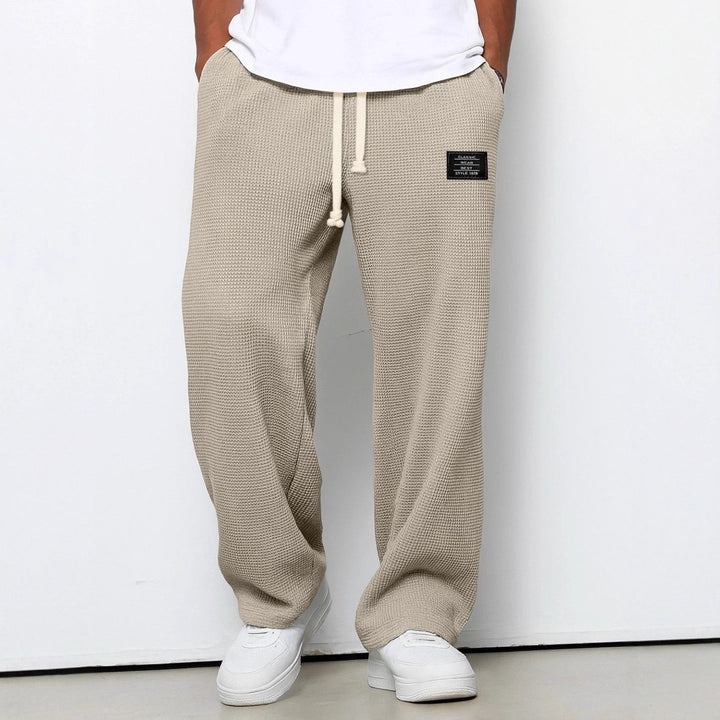 Beige joggers i ribstrik med snøre og sort mærke, bæres af person i hvid t-shirt og hvide sneakers. Komfortabelt og stilfuldt herretøj.
