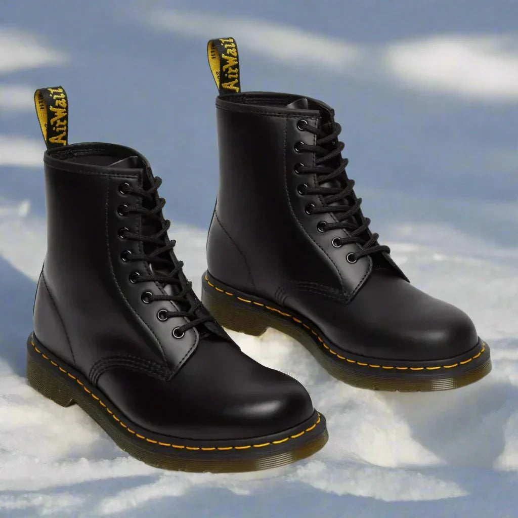 Sort Dr. Martens støvler i sneen, ikonisk design med gule syninger og snørebånd, robust læder, populært fodtøj til vinter og mode.