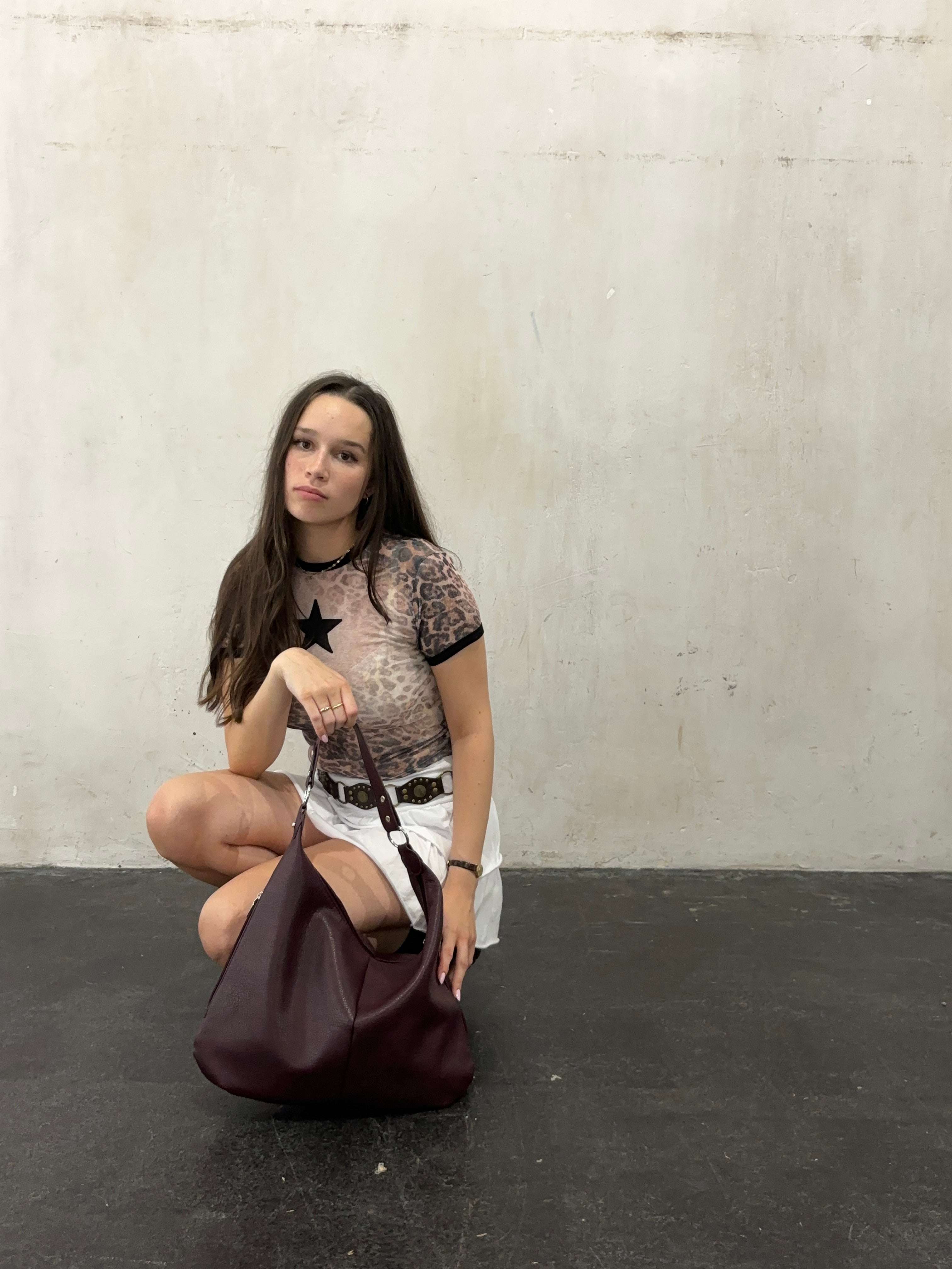 Ung kvinde i gennemsigtig bluse og hvid nederdel sidder på hug med en stor, bordeaux skuldertaske på mørkt gulv. Fashion, mode, stil.