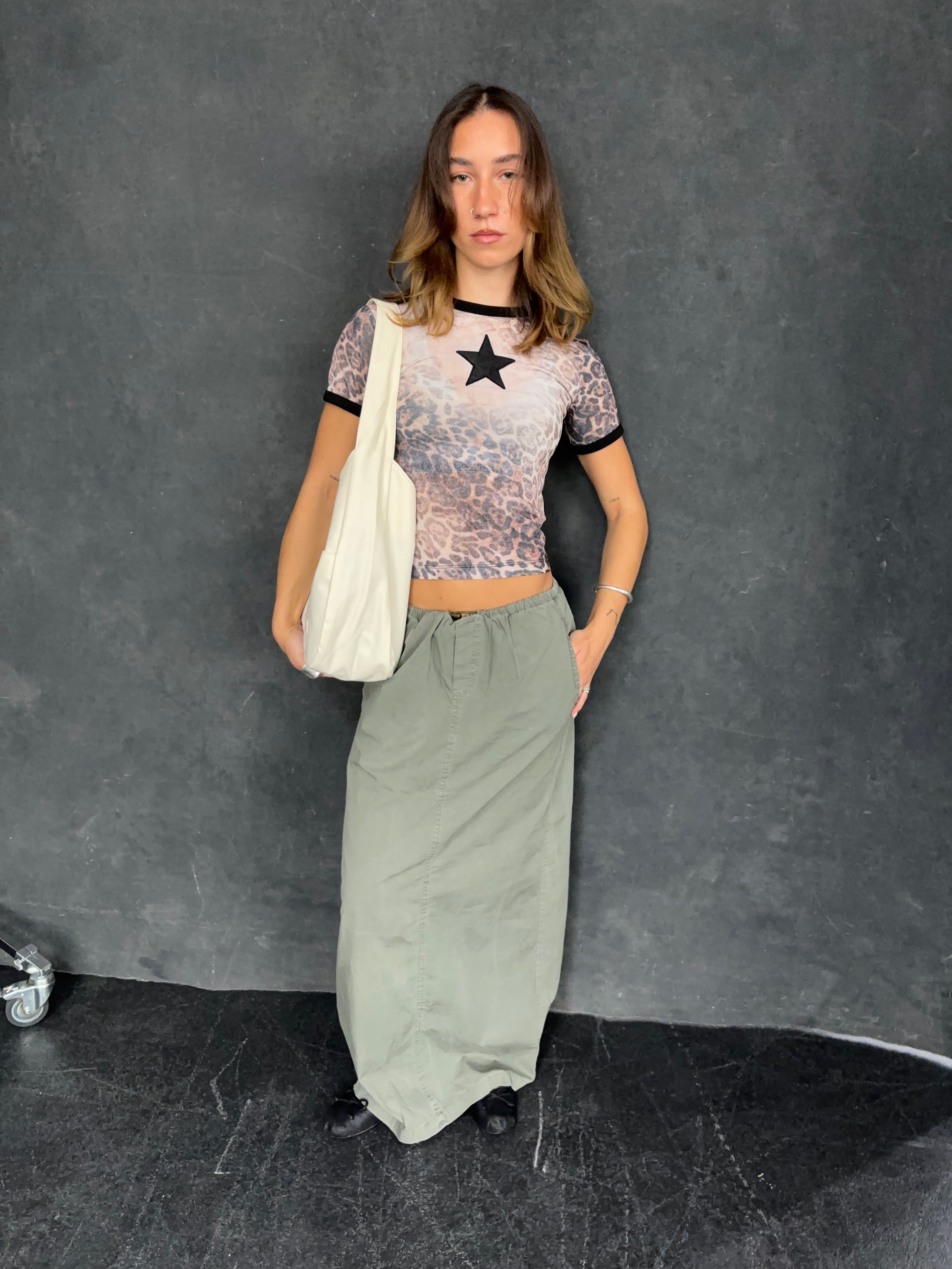 Ung kvinde i leopardmønstret top med stjerne, lang grøn nederdel og hvid taske. Mode, trendy tøj, casual stil, grå baggrund.