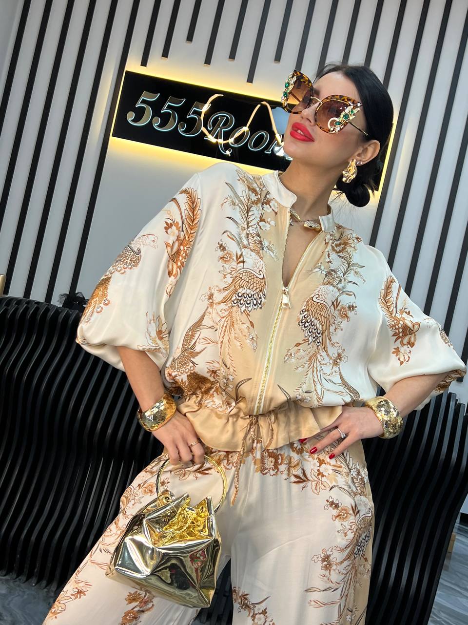 Kvinde i elegant, mønstret jumpsuit med store solbriller og guldtaske foran moderne 555Room skilt. Fashionabelt outfit, luksusmode, stilfuldt tilbehør.