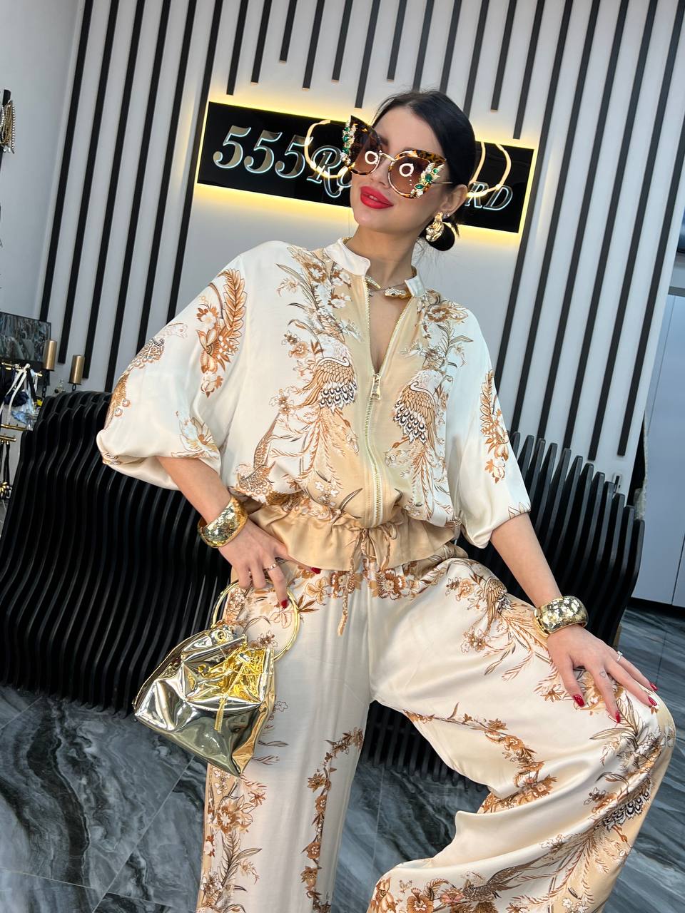 Kvinde i elegant, mønstret jumpsuit med gyldne detaljer, bærer en skinnende guldhåndtaske og solbriller. Moderne interiør i baggrunden.