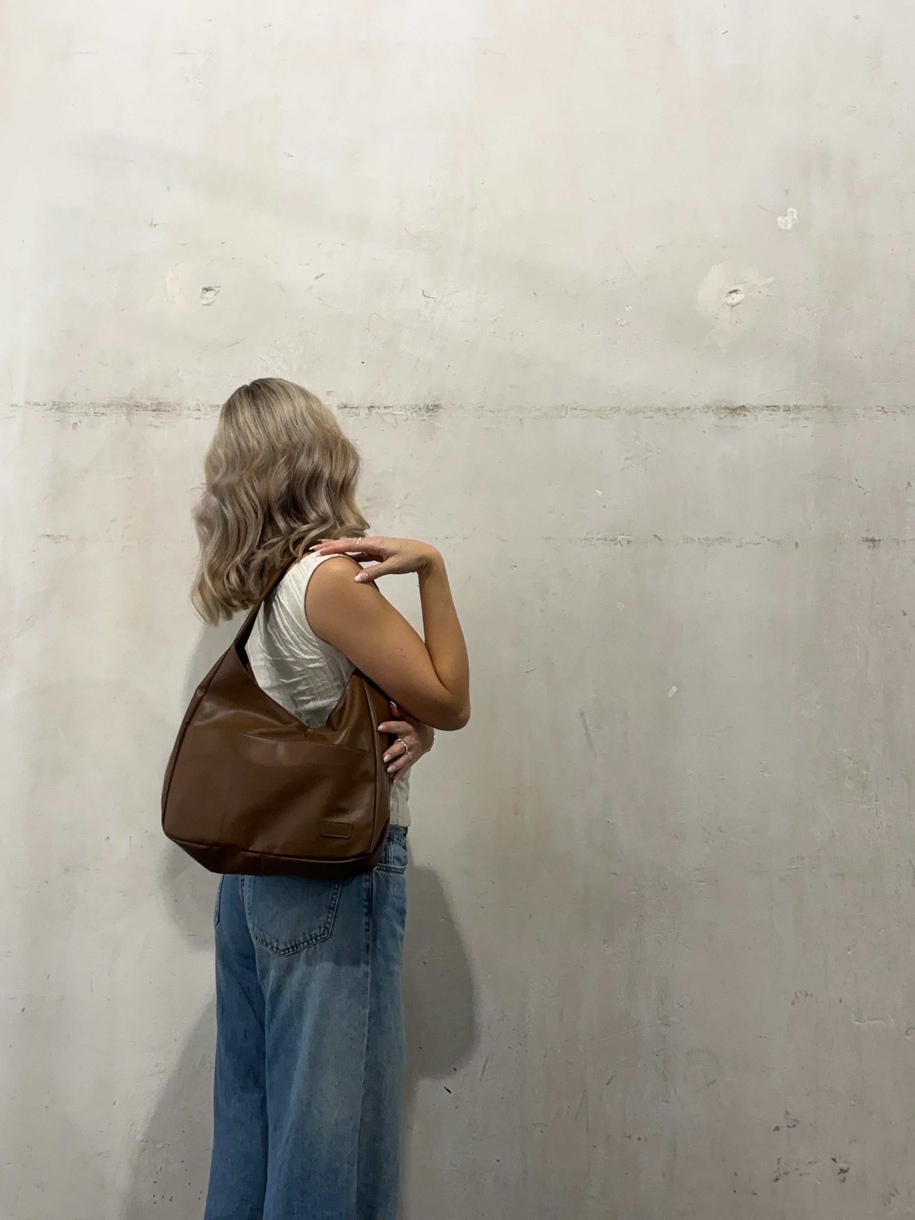 Kvinde med blondt hår iført hvid top og blå jeans, bærer en brun skuldertaske. Står mod en rå, hvid væg. Mode, casual stil, trendy tilbehør.