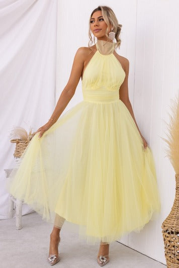Kvinde i gul tylkjole med halterneck, poserer elegant i lys indretning. Perfekt til sommerfest eller bryllup. Mode, kjole, kvinde, gul kjole.