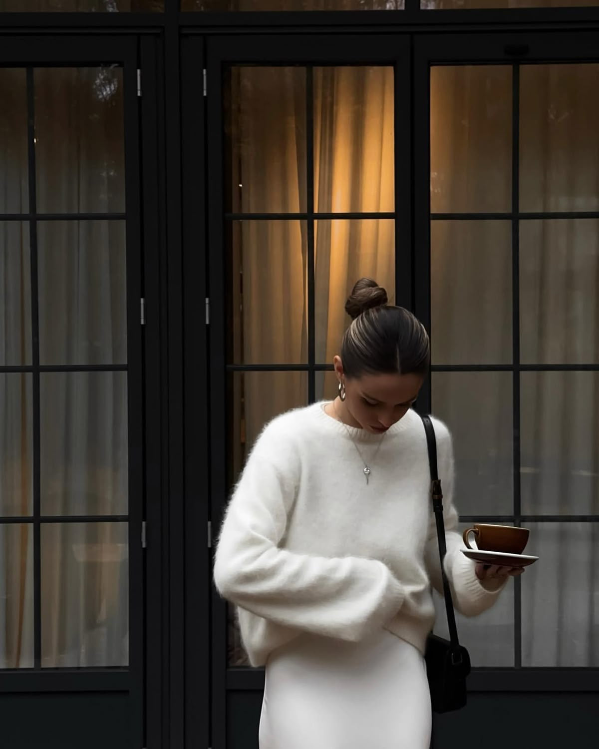 Kvinde i hvid sweater og nederdel foran glasdør, holder brun kaffekop. Elegant mode, minimalistisk stil, moderne kvindetøj, byliv.