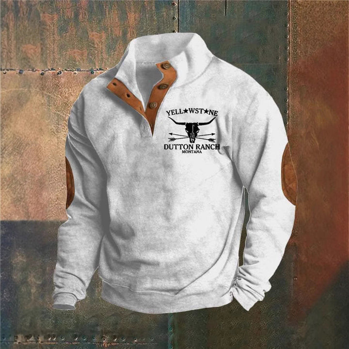 Hvid sweatshirt med brune albuelapper og knapper, "Yellowstone Dutton Ranch Montana" logo på brystet. Casual herretrøje, western stil.