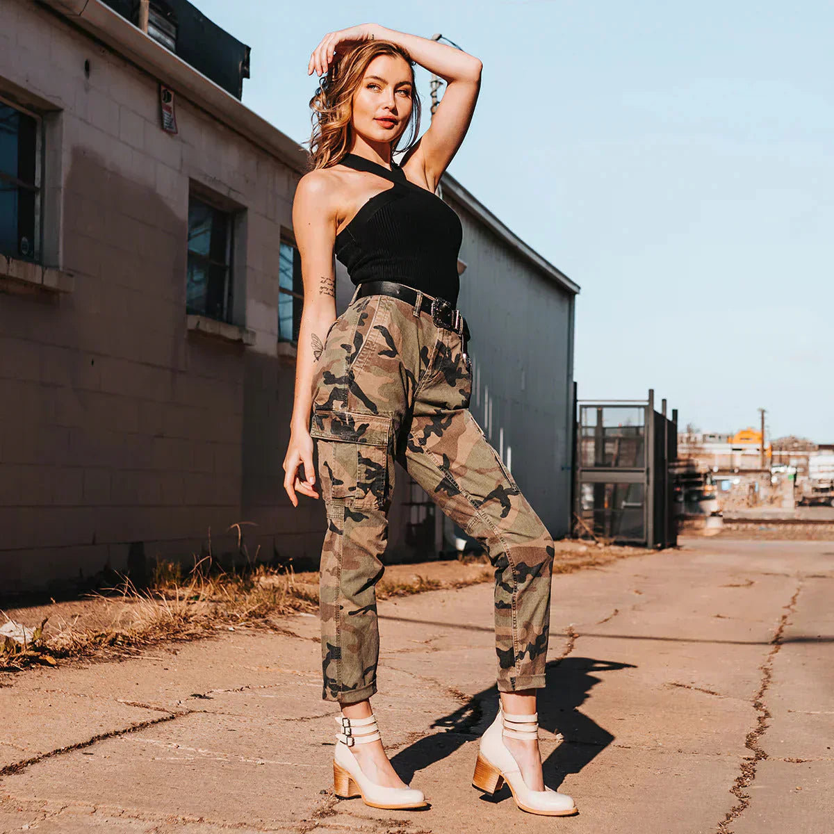 Kvinde i sort top og camouflagebukser poserer udendørs på en solrig dag. Mode, streetwear, stilfuldt outfit, moderne tøj, urban baggrund.