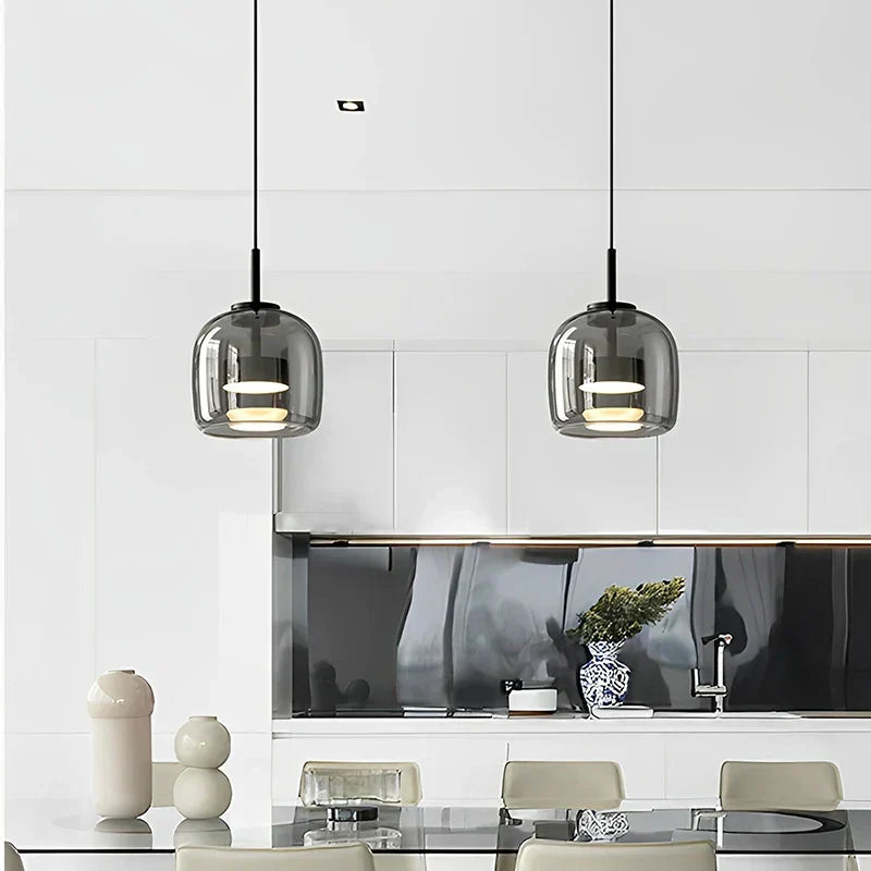To pendellamper i glas hænger over et moderne køkken med hvidt interiør og sort backsplash. Stilfuld indretning, minimalistisk design, skandinavisk stil.