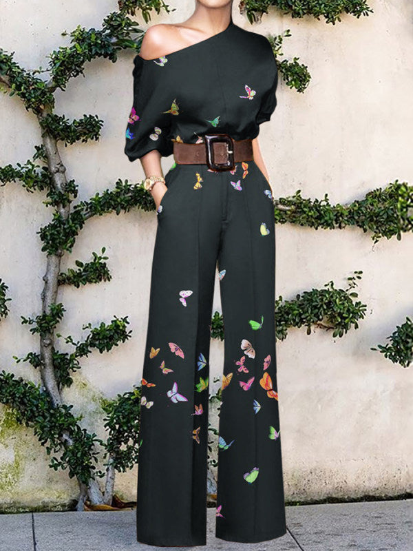 Kvinde i sort buksedragt med sommerfuglemønster, brun bælte, poserende foran væg med grønne planter. Elegant mode, trendy jumpsuit, sommerstil.