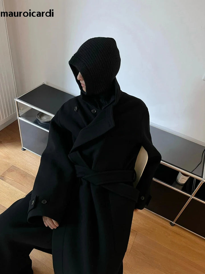 Sortklædt person i moderne, minimalistisk rum, iført sort frakke og hætte. Stilfuldt vinterlook, trendy mode, indendørs.