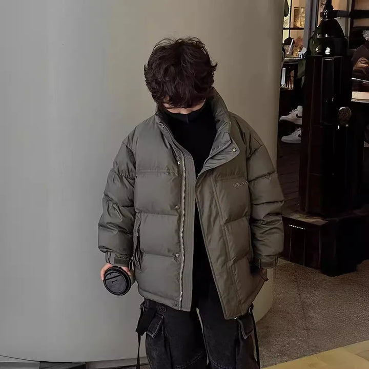 Dreng i grå dunjakke med hænderne i lommerne, holder en sort kop. Vintermode, børnetøj, varm jakke, indendørs.