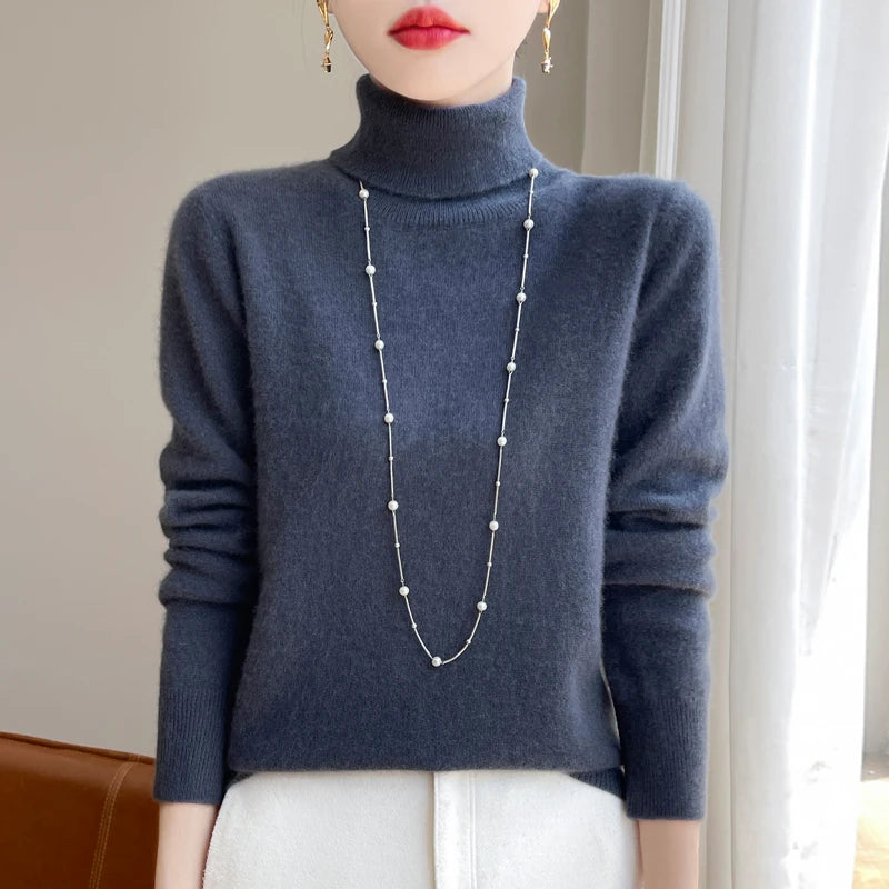 Kvinde iført grå rullekravetrøje og lang halskæde med perler, stående foran et vindue. Elegant vintermode, minimalistisk stil.