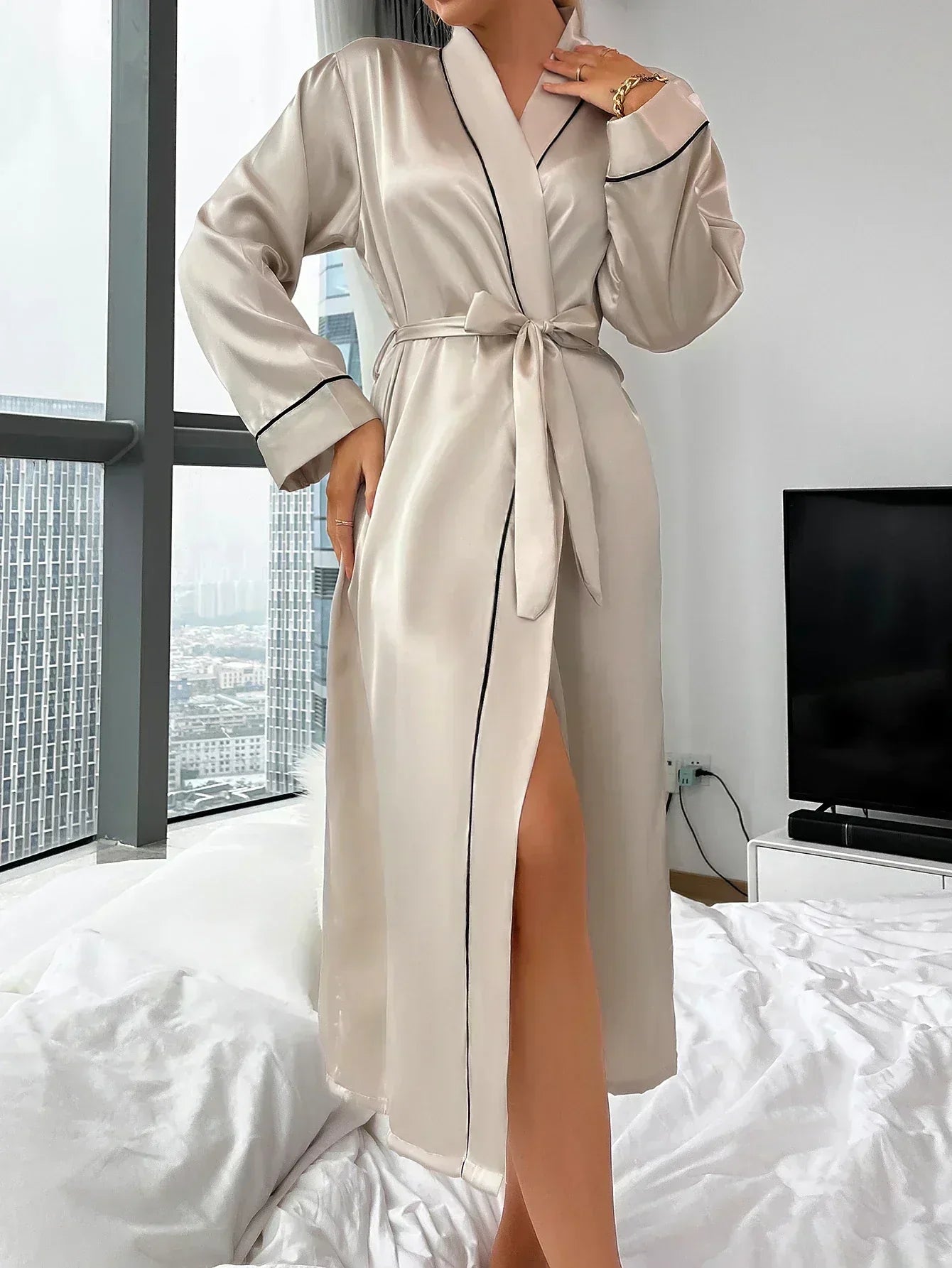 Kvinde iført elegant beige morgenkåbe med sort kant, står ved vindue i moderne soveværelse. Byudsigt i baggrunden. Luksus, komfort, mode.