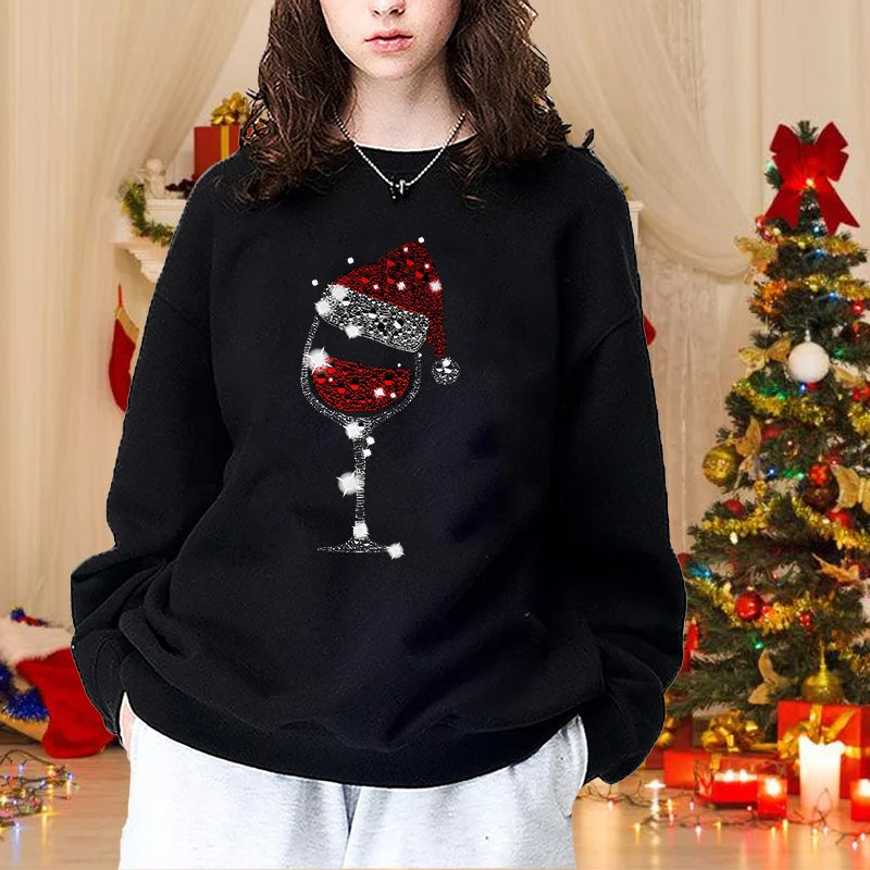 Ung kvinde i sort sweatshirt med julemotiv af vinglas og nissehue, står foran juletræ. Perfekt til julefest, vintermode, hyggestil.