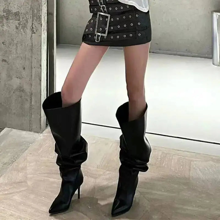 Kvinde iført sorte, høje støvler med spidse tæer og en sort nederdel med bælte. Moderne mode, stilfuldt outfit, trendy fodtøj.