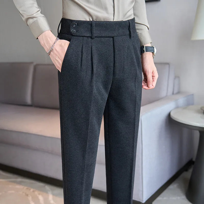 Mand iført grå bukser med høj talje og knapdetaljer, stående i moderne stue. Elegant herremode, klassisk design, stilfuldt outfit.