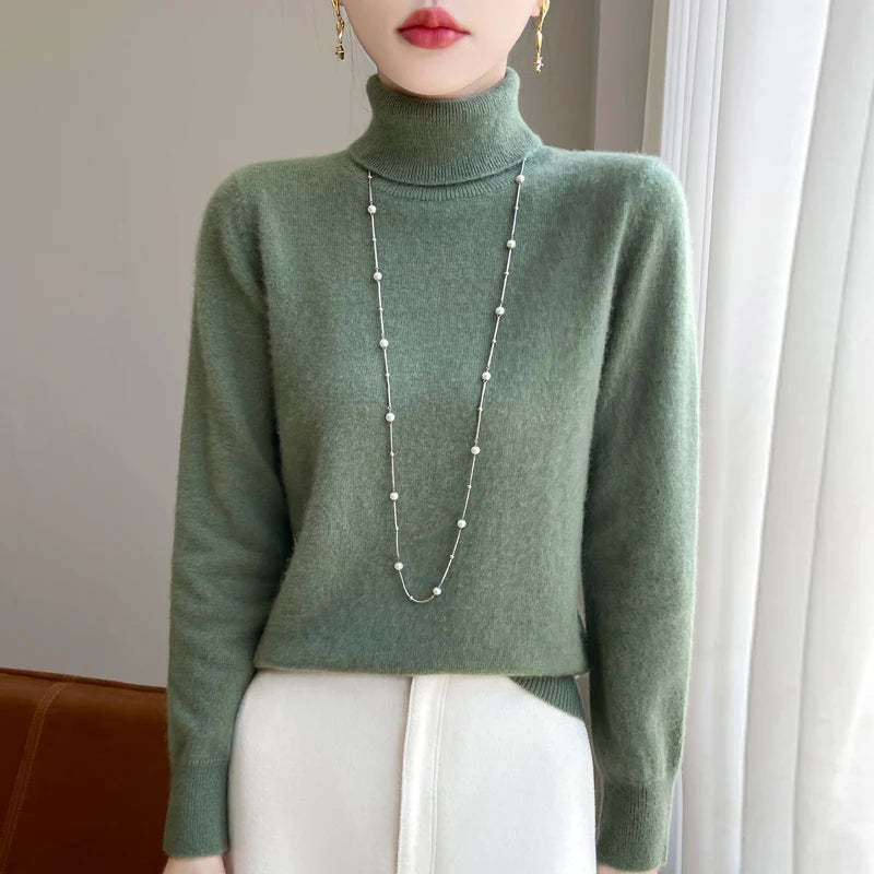 Kvinde iført grøn rullekrave sweater og lang halskæde, stående foran hvidt gardin. Elegant vintermode, minimalistisk stil, moderne kvindetøj.