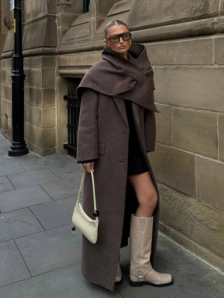 Kvinde i brun frakke og beige støvler står op ad murstensvæg, bærer hvid taske. Mode, vintertøj, stilfuldt look, bymiljø.