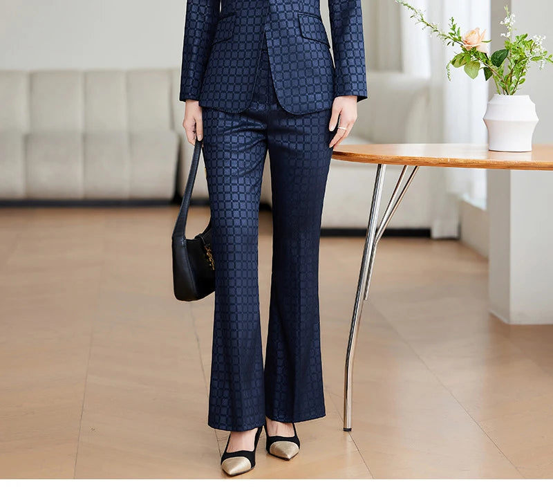 Kvinde i elegant, mørkeblå jakkesæt med mønster, står ved træbord med vase og blomster. Moderne kontorindretning. Businessmode, stilfuldt look.