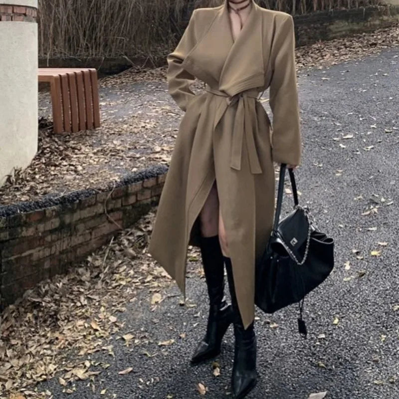 Kvinde iført beige frakke med bælte, sorte knæhøje støvler og sort taske på en bladfyldt sti. Elegant efterårsoutfit, mode, udendørs.