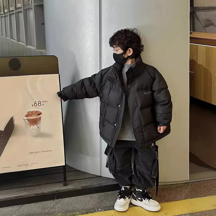 Dreng i sort vinterjakke og maske peger på caféplakat med kaffe. Moderne børnemode, vintertøj, cafébesøg.