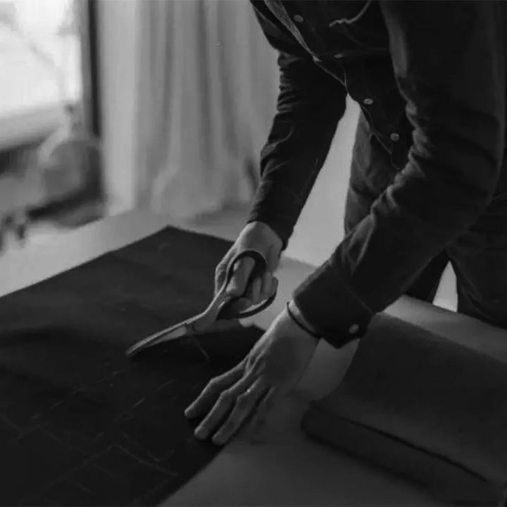 Person skærer stof med saks på arbejdsbord. Skrædderi, håndværk, tekstilarbejde. Sort-hvid billede af præcisionsarbejde i modeindustrien.