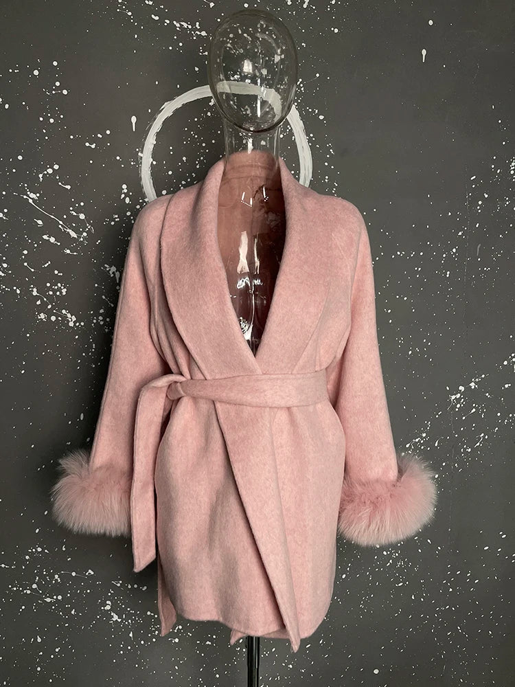 Lyserød uldfrakke med bælte og pelsdetaljer på ærmerne, udstillet på gennemsigtig mannequin mod mørk, stænket baggrund.