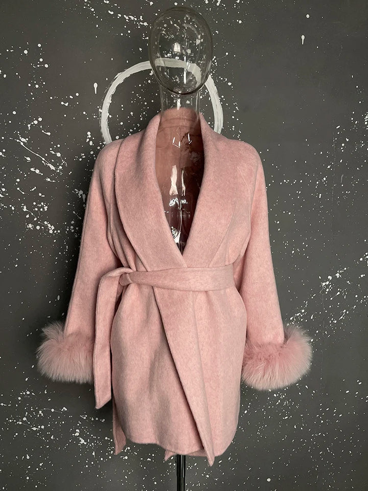 Lyserød uldfrakke med bælte og pelsdetaljer på ærmerne, udstillet på gennemsigtig mannequin mod mørk, stænket baggrund.