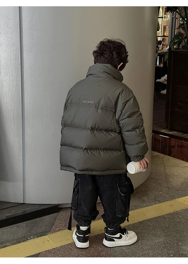 Lille barn iført grå dunjakke og sorte bukser med sneakers, holder en hvid flaske. Børnemode, vintertøj, udendørs.