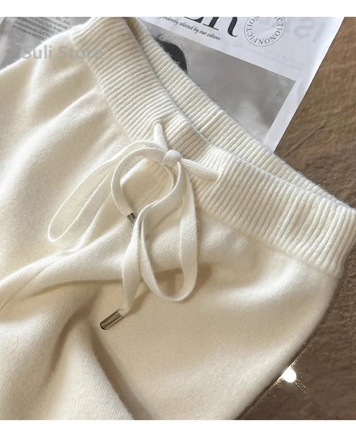 Hvide strikbukser med snøre i taljen, liggende på et bord med en avis i baggrunden. Komfortable og stilfulde bukser til afslappet brug.