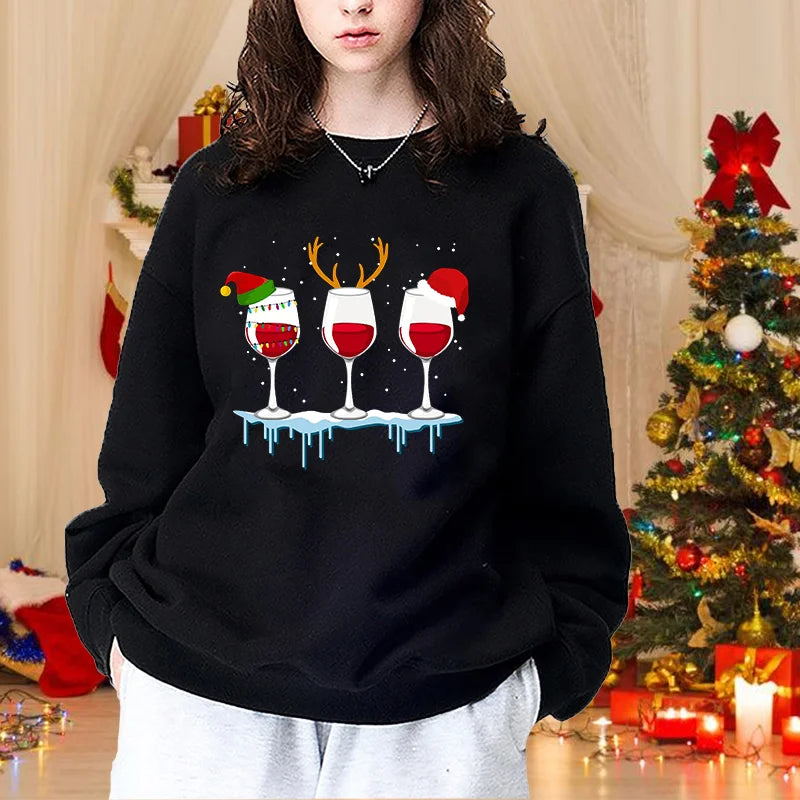 Kvinde i sort sweatshirt med julemotiv af tre vinglas, står foran juletræ. Perfekt til julefest, vintermode, hyggelig julestemning.