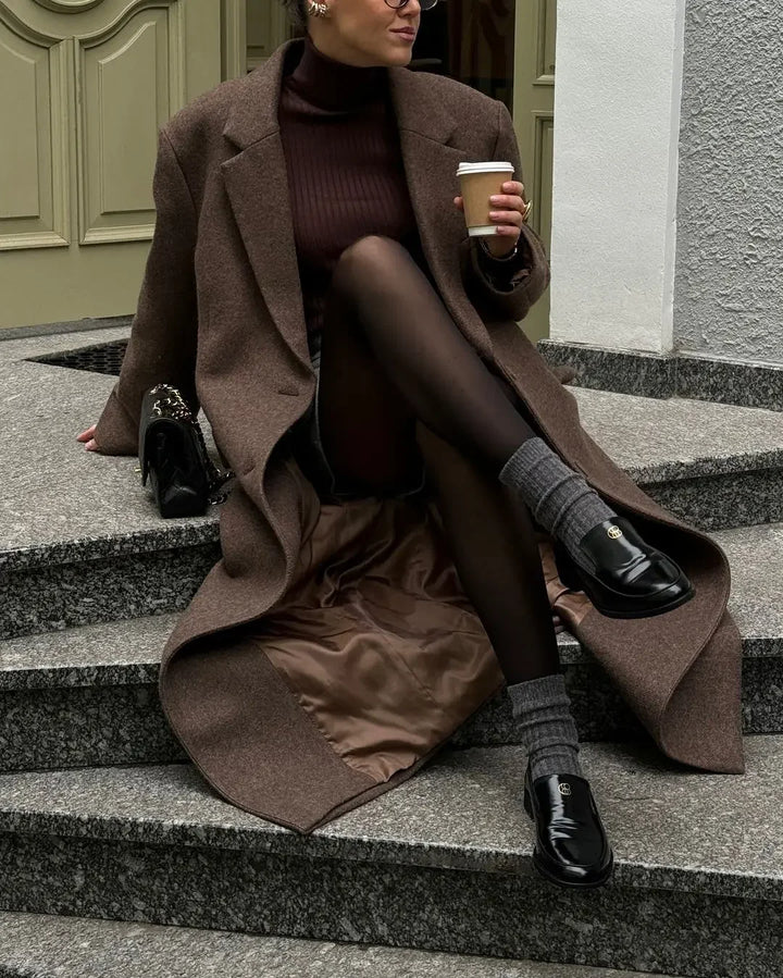 Kvinde i brun frakke og sorte loafers sidder på trappe med kaffekop. Elegant efterårsoutfit, mode, stilfuldt tøj, byliv.