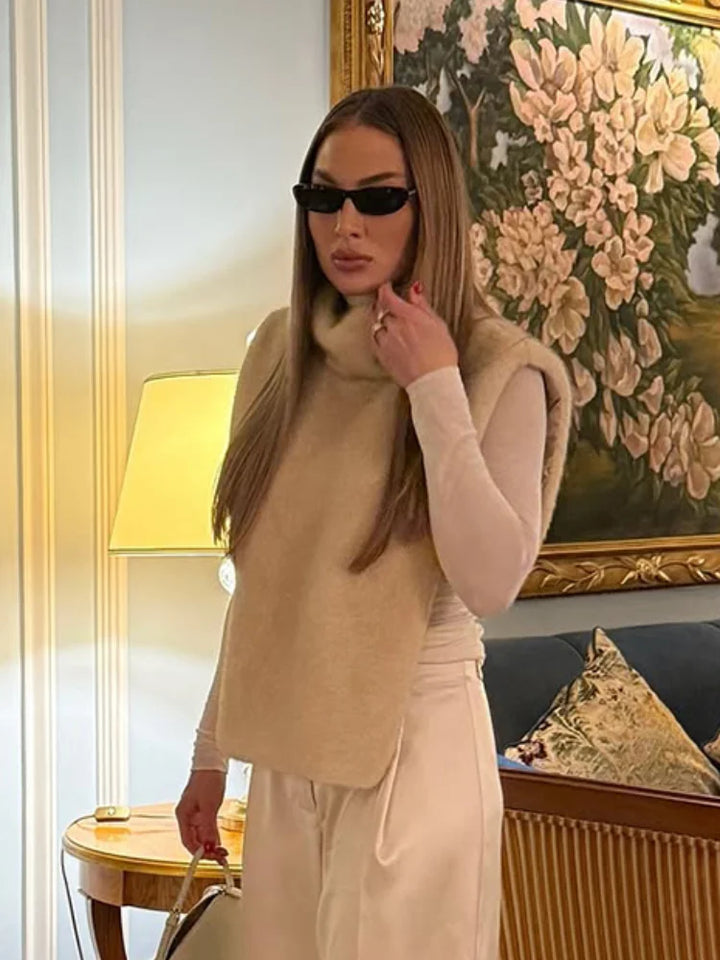 Kvinde med langt hår iført solbriller og beige tøj står i elegant stue med blomsterbillede og gulvlampe. Luksuriøs indretning, moderne mode.