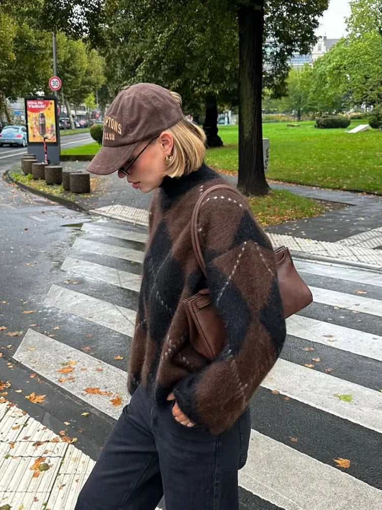 Kvinde i brunt og sort ternet sweater og kasket krydser fodgængerfelt i bypark. Efterårsdag med grønne træer og våde gader.