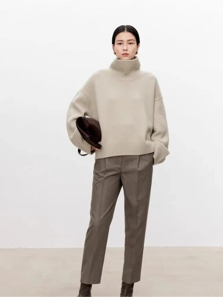 Kvinde iført beige rullekravesweater og grå bukser, holder brun hat. Minimalistisk mode, efterårsoutfit, stilfuldt og moderne look.