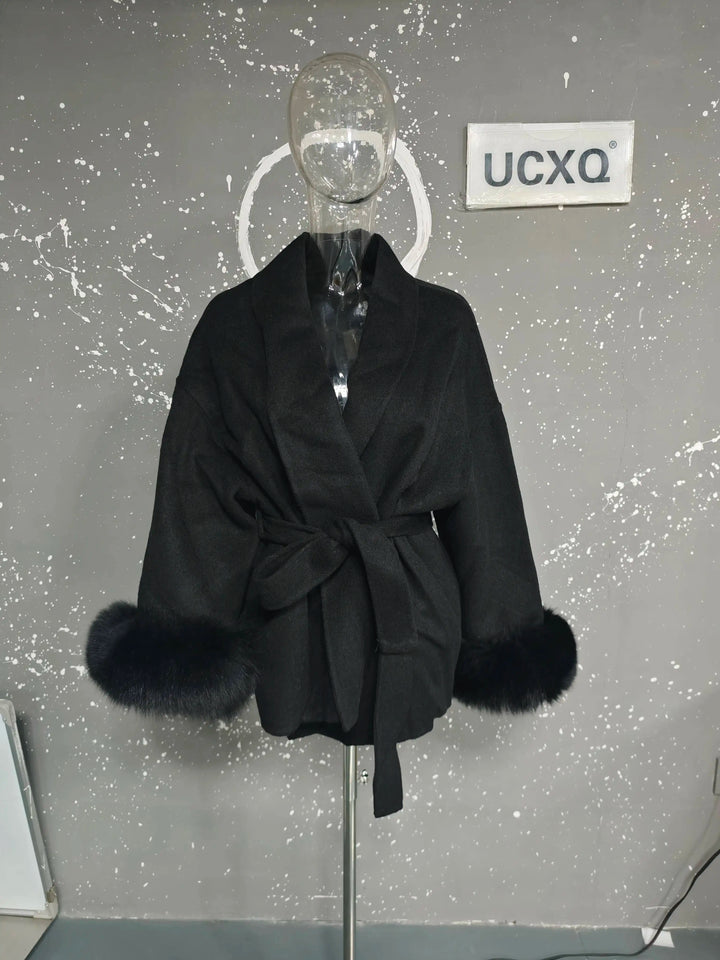 Sort frakke med bælte og pelsmanchetter på mannequin mod grå stænket væg. Elegant vinterjakke, mode, kvindetøj, stilfuldt design.