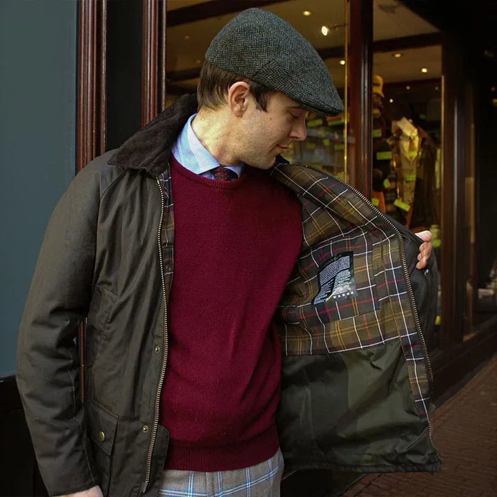 Mand iført brun jakke med ternet for, rød sweater og flad kasket foran butik. Klassisk herremode, efterårsjakke, stilfuldt tøjvalg.