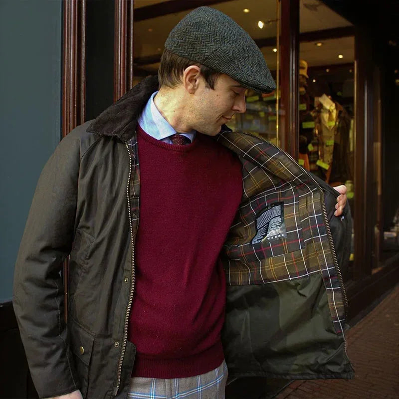 Mand iført brun jakke med ternet for, rød sweater og flad kasket foran butik. Klassisk herremode, efterårsjakke, stilfuldt tøjvalg.