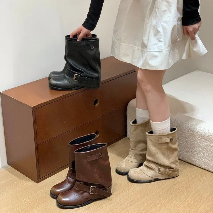 Kvinde iført hvid kjole og sorte sokker, vælger mellem tre par støvler i sort, brun og beige på trækommode. Mode, fodtøj, stil.