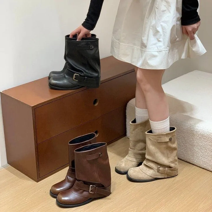 Kvinde iført hvid kjole og sorte sokker, vælger mellem tre par støvler i sort, brun og beige på trækommode. Mode, fodtøj, stil.