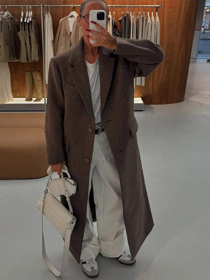 Kvinde i spejlselfie iført lang brun frakke, hvid T-shirt og hvide bukser, med hvid taske og sneakers, i moderne tøjbutik.