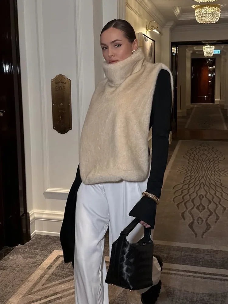 Kvinde i moderne tøj med beige vest og sorte ærmer, står i elegant hotelgang. Hun bærer en sort taske. Luksuriøs mode, stilfuldt outfit.