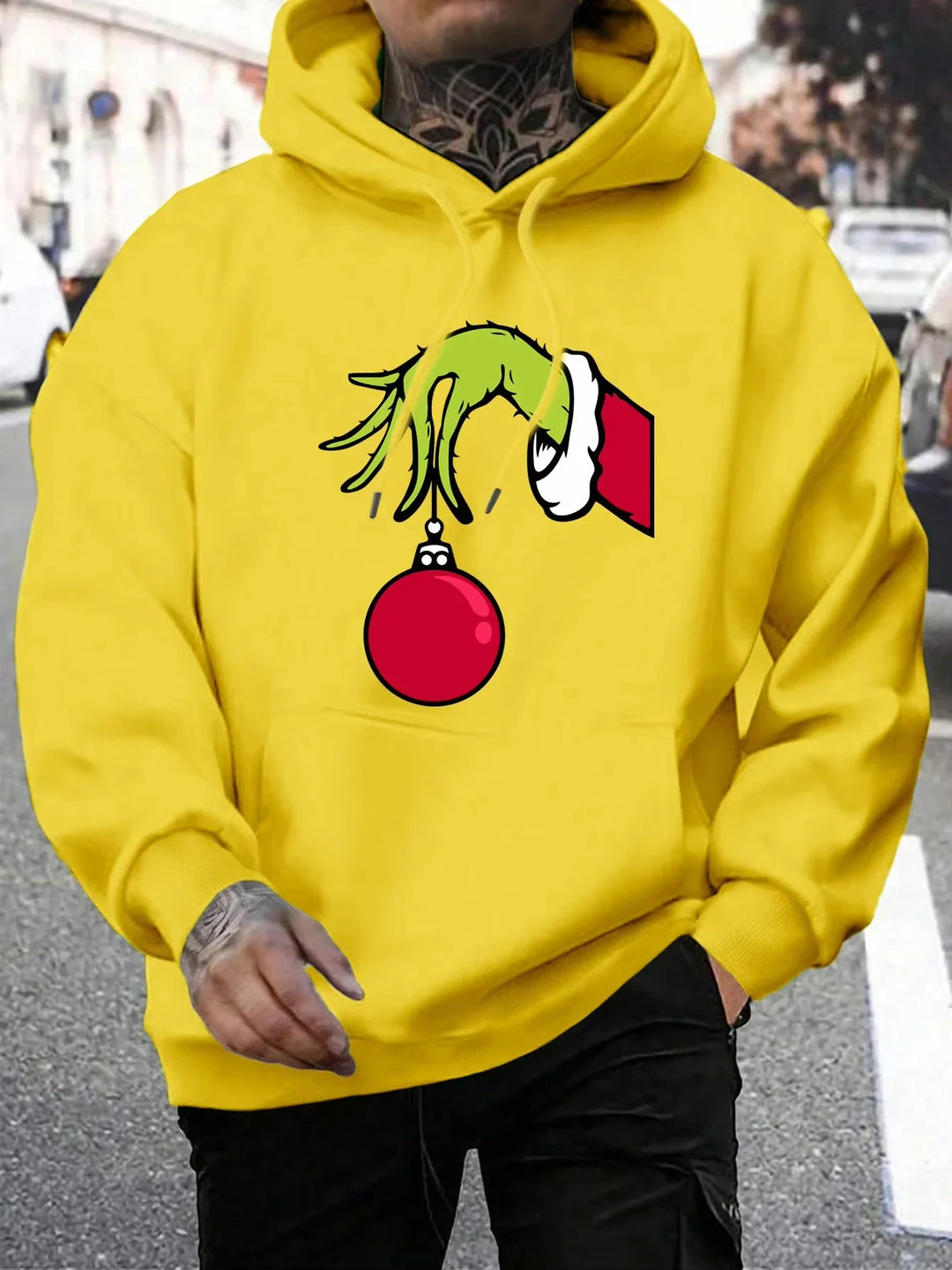 Mand iført gul hættetrøje med Grinch-motiv, der holder en rød julekugle. Streetwear, julemode, unisex hoodie, trendy design.