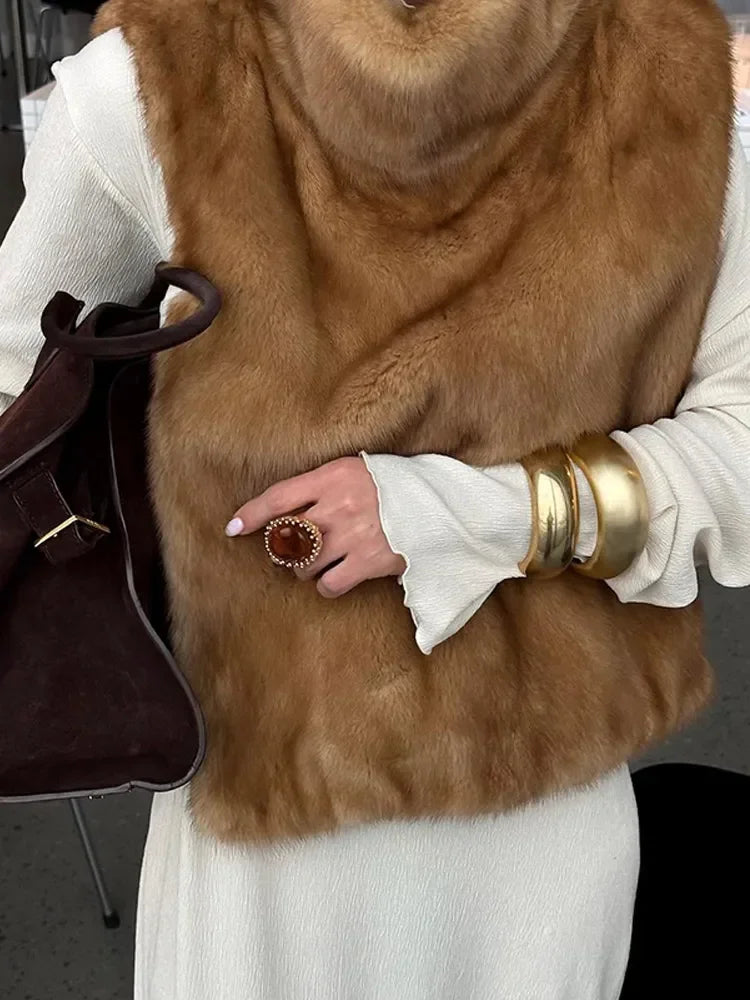 Kvinde iført hvid kjole og brun pelsvest, bærer brun taske. Guldarmbånd og stor ring på fingeren. Elegant mode, luksus accessories.