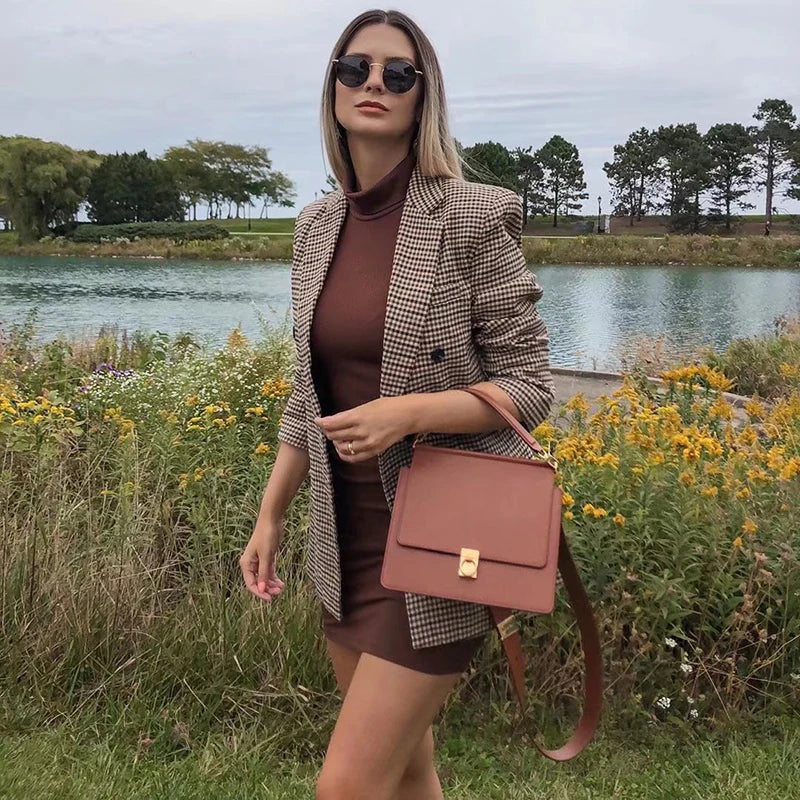 Kvinde i ternet blazer og brun kjole ved sø, bærer brun taske. Solbriller, naturbaggrund med blomster og træer. Mode, efterår, stil.